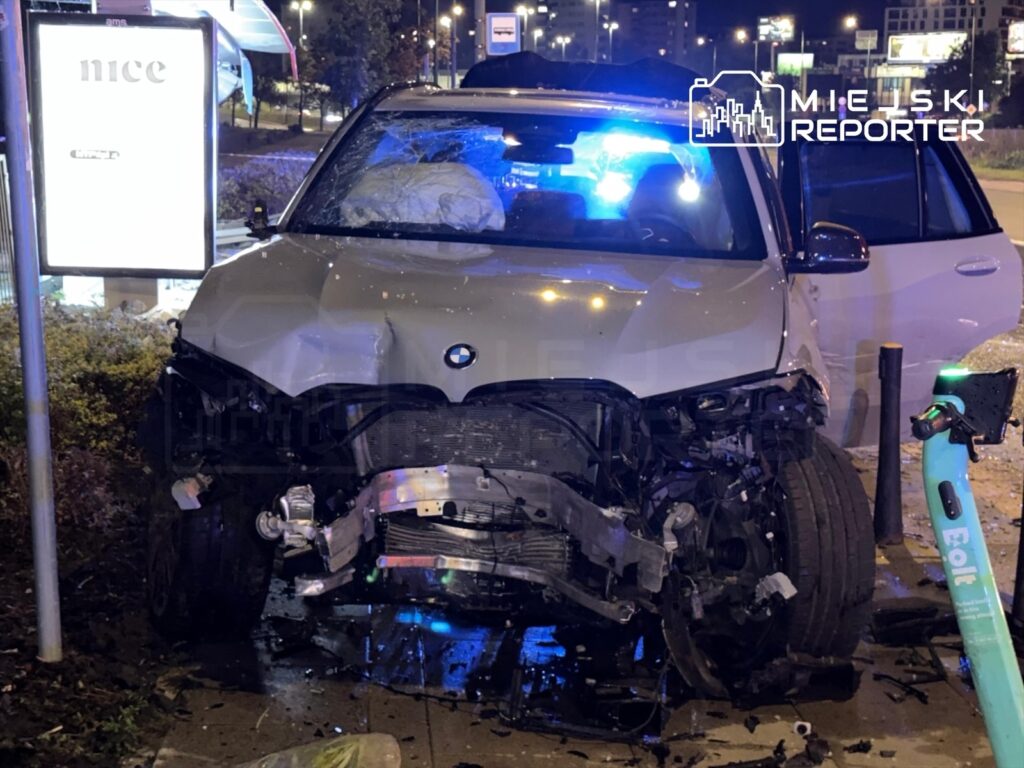 BMW z uszkodzoną przednią częścią stoi obok przystanku, z włączonymi niebieskimi światłami policyjnymi w tle.