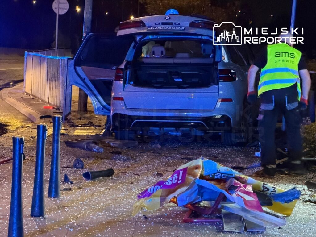 Samochód osobowy z otwartymi drzwiami stoi obok uszkodzonego przystanku, a pracownik w odblaskowej kamizelce zbiera rozbite elementy.