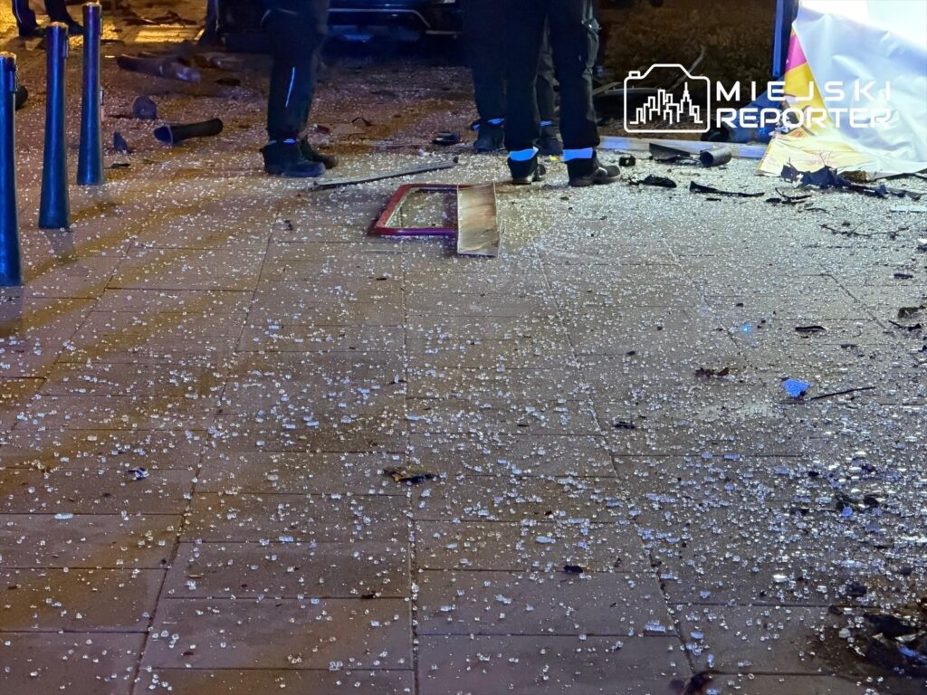 Na chodniku leżą odłamki szkła oraz resztki zniszczonego przystanku, obok stoją ratownicy.