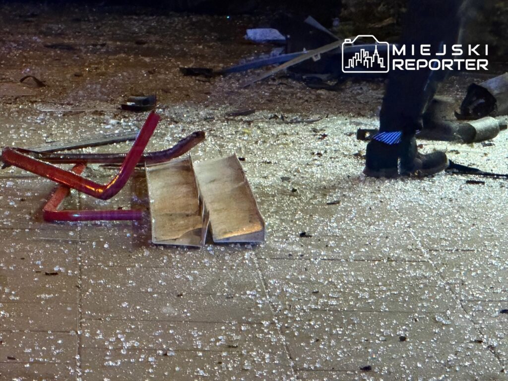 Szkło rozbite na chodniku, obok leżą metalowe elementy i fragmenty mebli. W tle widoczna stopa osoby w czarnych butach.