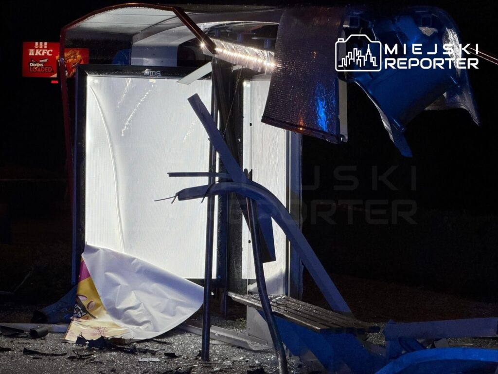Zniszczony przystanek autobusowy z uszkodzonym dachem i porozrzucanymi elementami, w tle neonowe światło KFC.