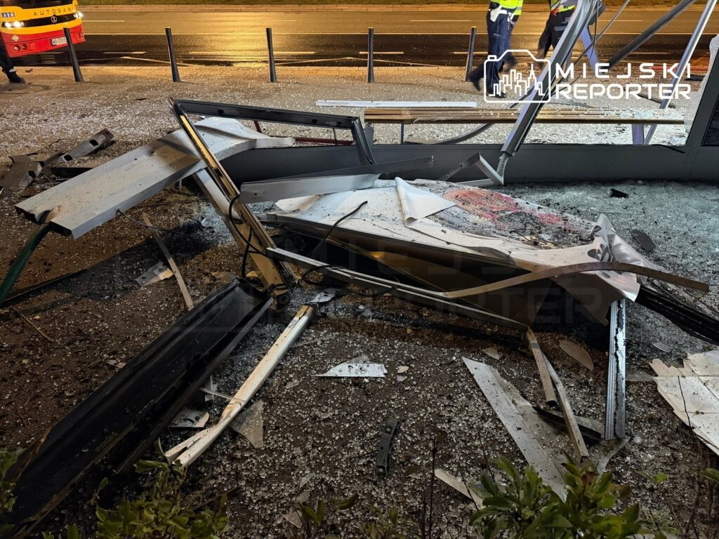Zniszczony przystanek autobusowy z porozrzucanymi fragmentami szkła i metalowymi elementami na jezdni.