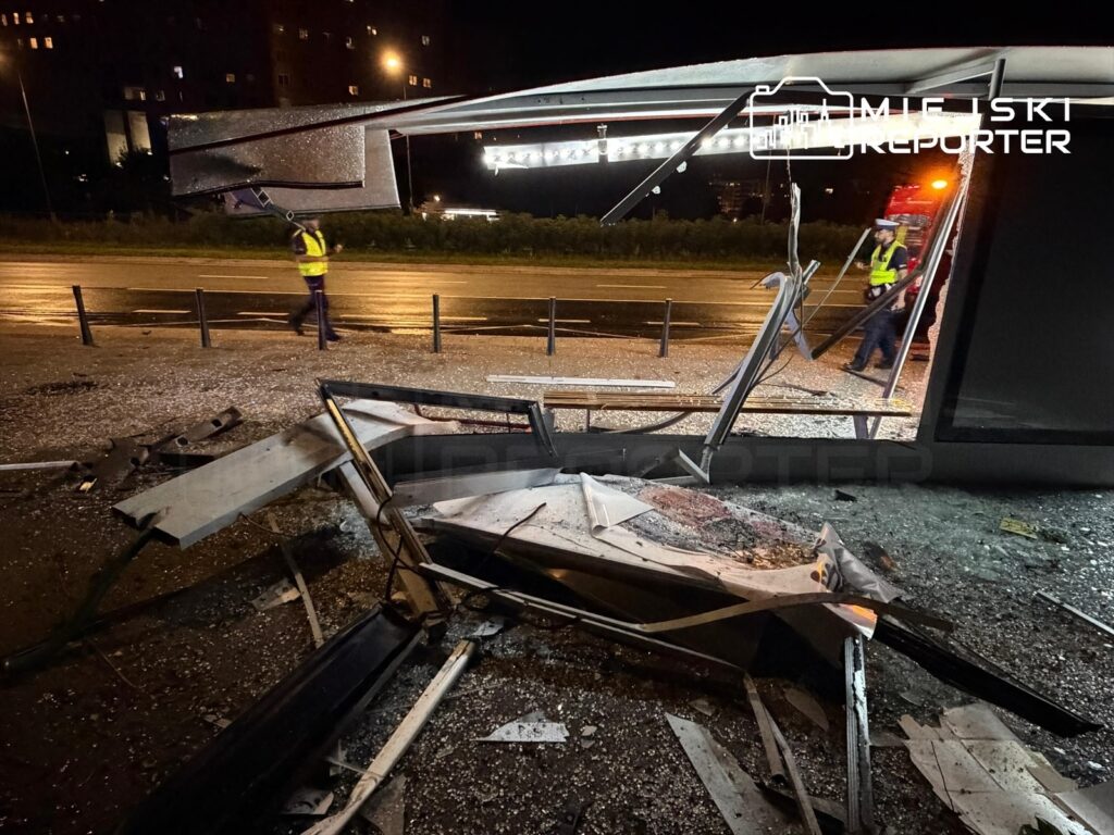 Zniszczony przystanek autobusowy z rozbitym szkłem i metalowymi elementami, obok pracujący ratownicy w odblaskowych kamizelkach.