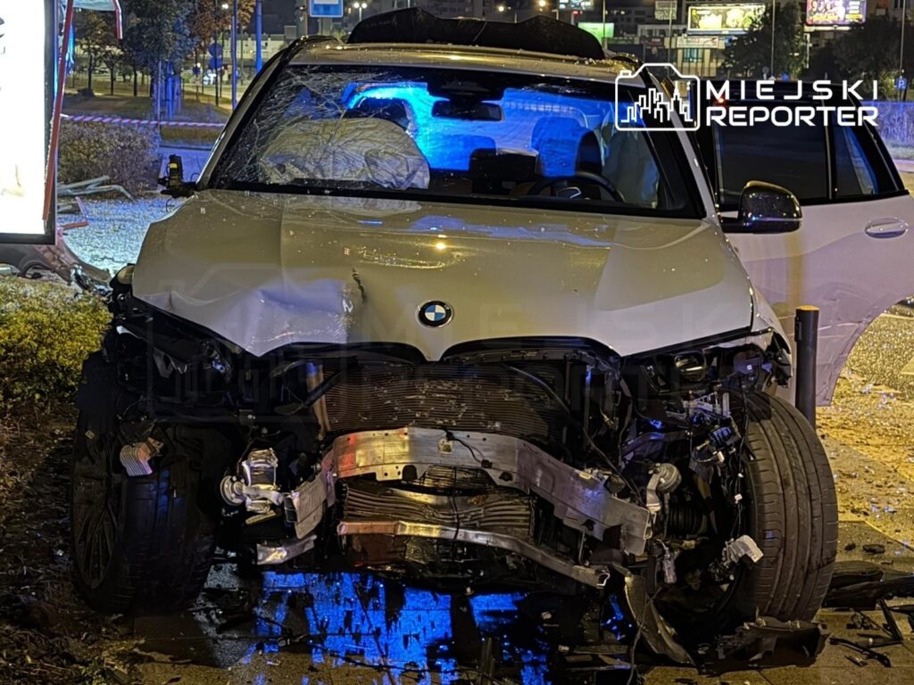 Zniszczone BMW z uszkodzoną przednią częścią stoi na chodniku obok przystanku, oświetlone niebieskimi światłami.