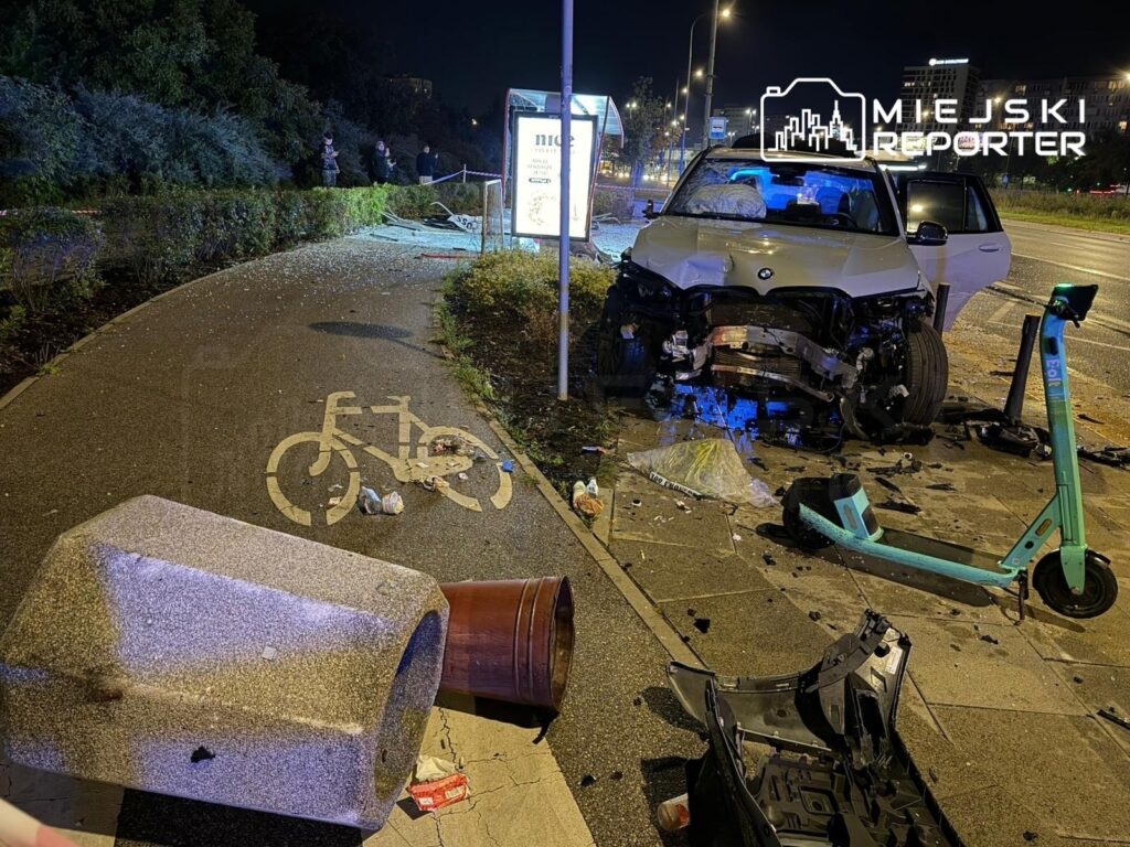 BMW z uszkodzonym przodem stoi na chodniku, obok przewrócony kosz na śmieci oraz rozbite elementy otoczenia.