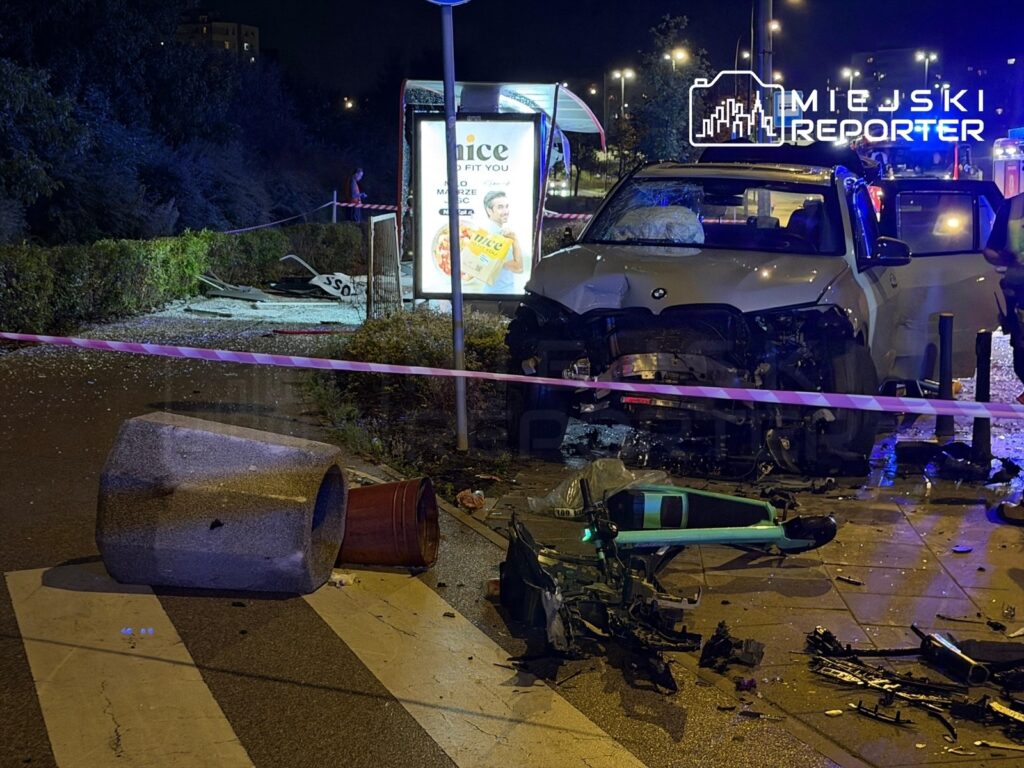BMW z uszkodzoną przednią częścią stoi przy przystanku, obok leżą zniszczone elementy infrastruktury oraz połamane donice.