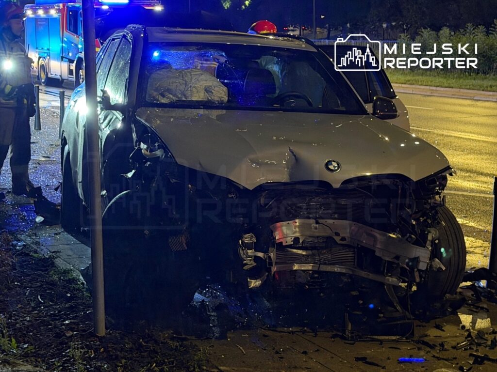 Zniszczony samochód marki BMW uderzył w słup przystanku, obok pracują służby ratunkowe w nocy.