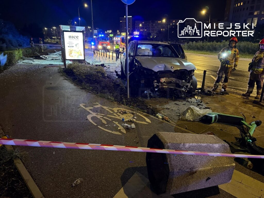 BMW z uszkodzoną przednią częścią stoi na chodniku obok zniszczonego przystanku, a w tle widać pojazdy ratunkowe.
