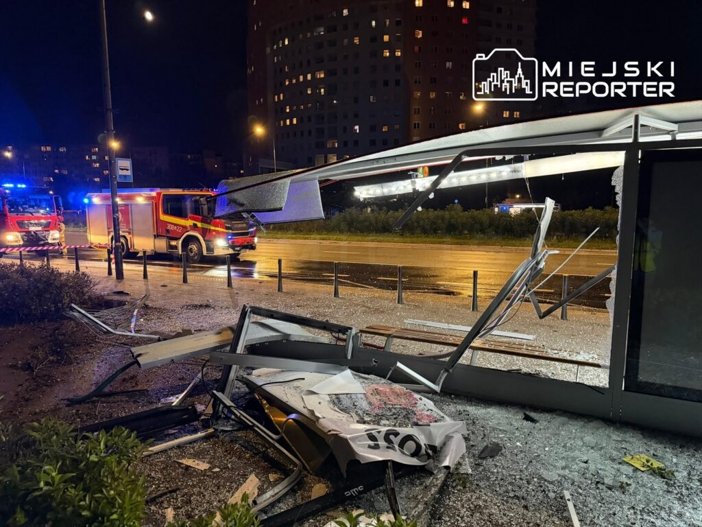 Zniszczony przystanek autobusowy z rozbitym szkłem i uszkodzoną konstrukcją, w tle woz strażacki.