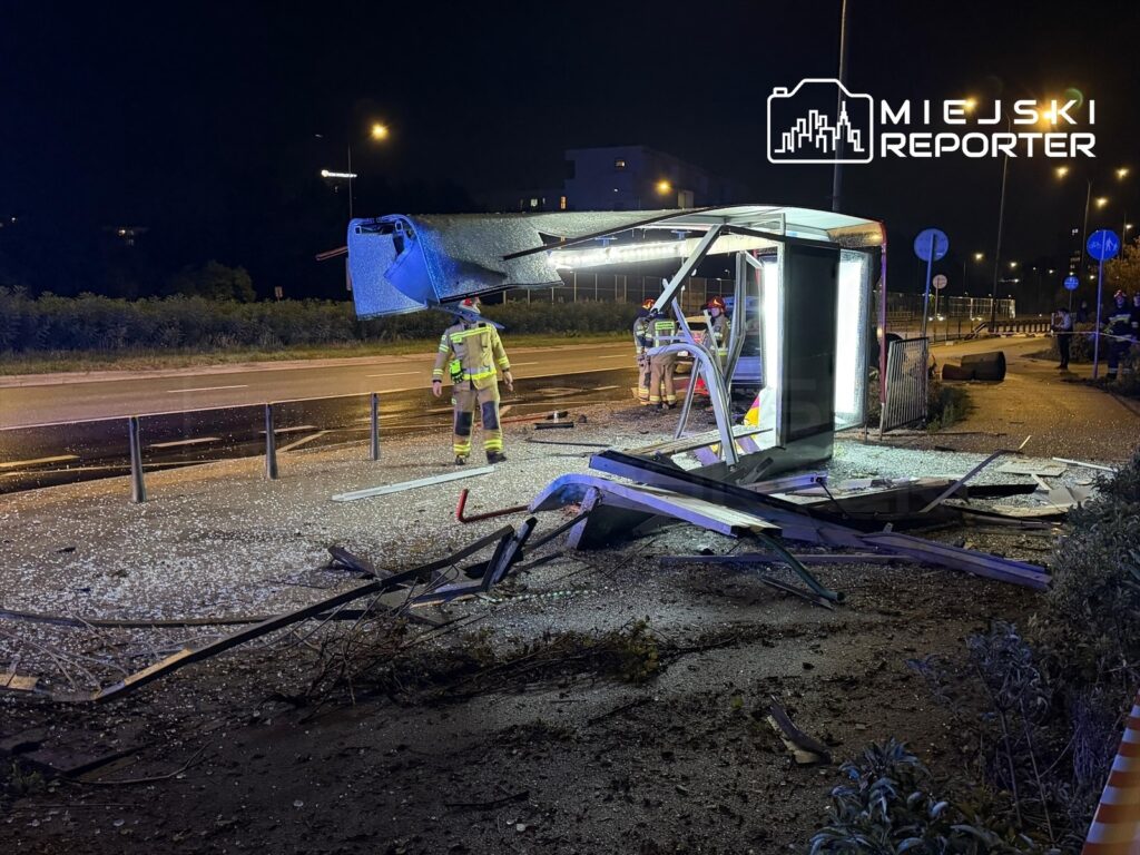 Strażacy zabezpieczają uszkodzony przystanek autobusowy po wypadku, na którym leżą odłamki i zniszczone elementy.