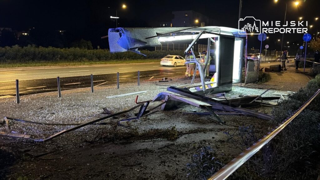 Zniszczony przystanek autobusowy z uszkodzonym zadaszeniem i porozrzucanymi elementami na jezdni po wypadku.