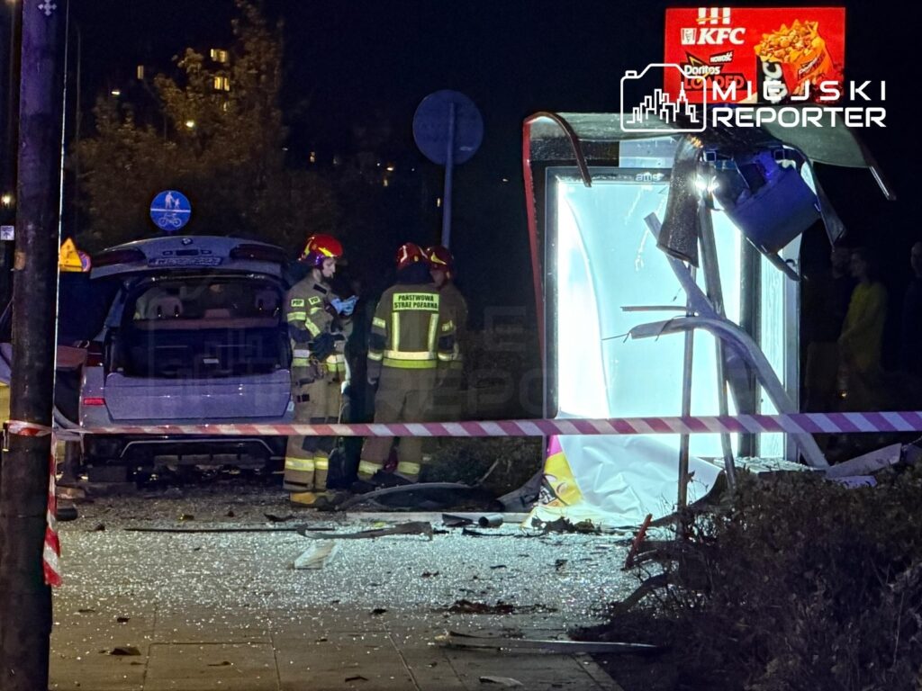 Strażacy w hełmach badają wypadek, w którym samochód uderzył w przystanek, a wokół leżą odłamki szkła.