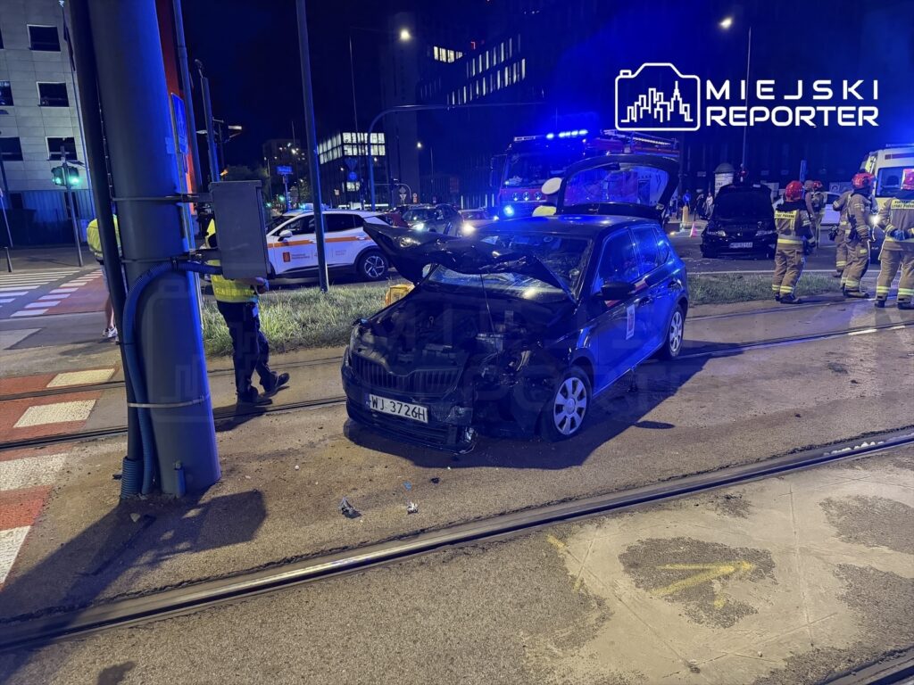 Czarny samochód osobowy z uszkodzoną maską stoi obok słupa trakcyjnego, wokół pracują strażacy i policja.