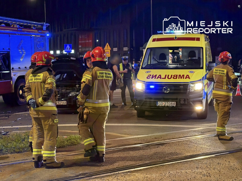 Grupa strażaków w mundurach omawia sytuację przy uszkodzonej Skodzie, obok stoi ambulans.