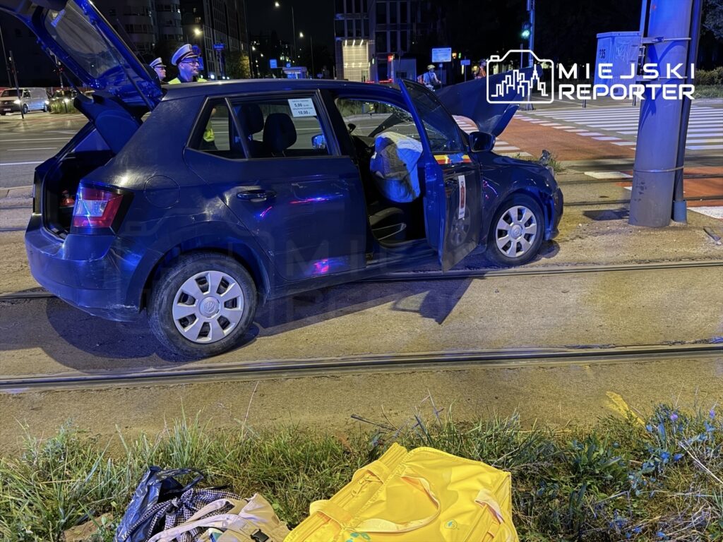 Czarny samochód osobowy z otwartą klapą bagażnika stoi na torach tramwajowych, obok dwóch policjantów w mundurach.