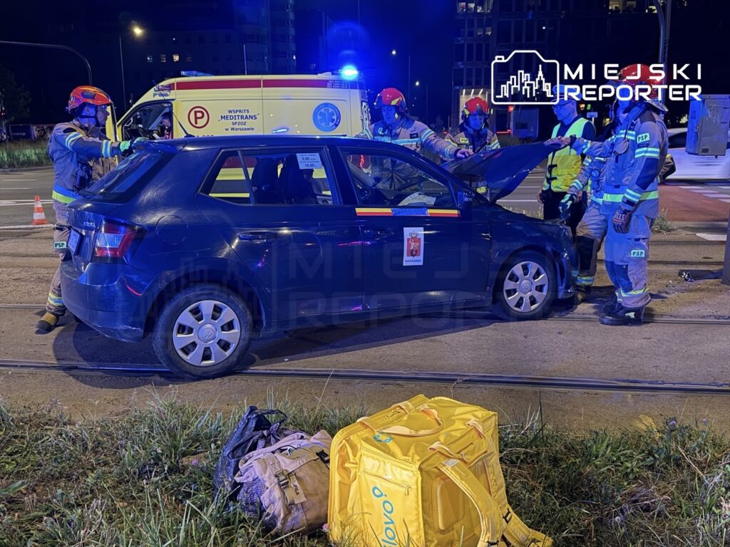 Ratownicy medyczni sprawdzają uszkodzone auto, które uderzyło w słup trakcyjny, na ulicy w nocy. W tle widoczna karetka.