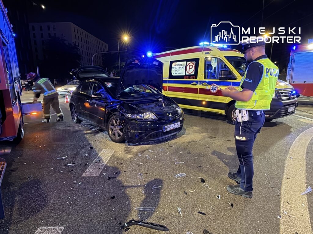 Czarny samochód osobowy z uszkodzoną maską stoi na drodze, obok policjant notujący szczegóły zdarzenia, w tle wozem strażackim.