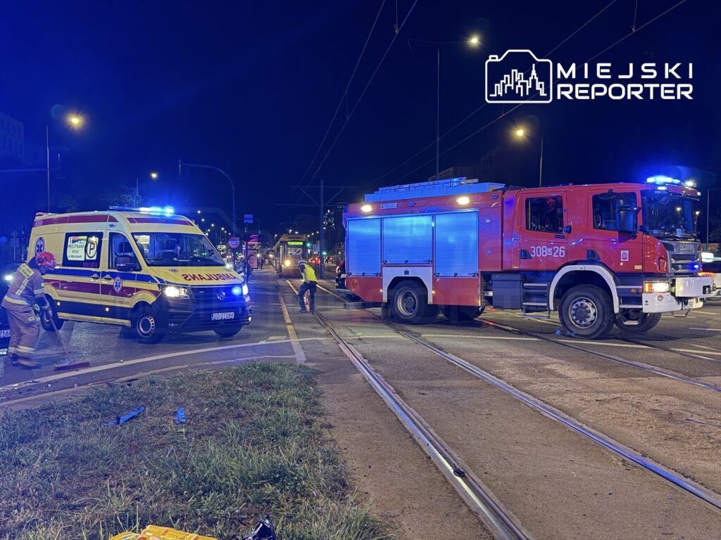 Ambulans i wóz strażacki z włączonymi sygnałami na ulicy, gdzie strażacy sprzątają po wypadku drogowym.