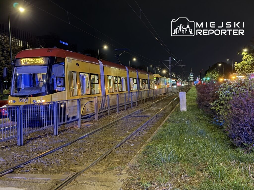 Tramwaj linii 17 zatrzymany na torowisku, oświetlony nocnymi lampami, w tle widać zarys miasta z kolorowymi światłami.