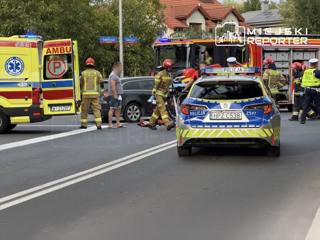 Na ulicy interweniują strażacy i policjanci, pomagając poszkodowanemu motocykliście po wypadku.