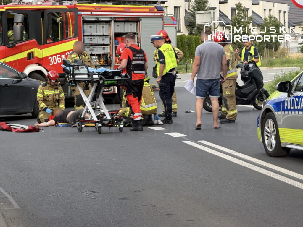Grupa ratowników medycznych udziela pomocy motocykliście leżącemu na jezdni obok wozu strażackiego i radiowozu policji.