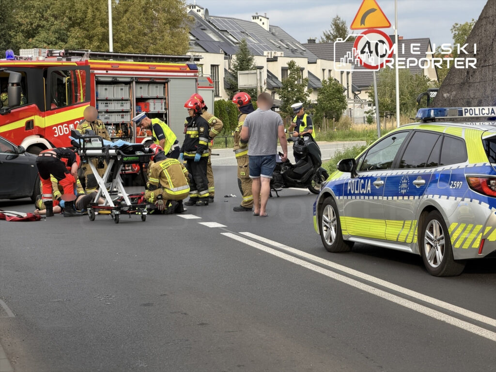 Grupa ratowników medycznych i strażaków udziela pomocy motocykliście na jezdni, obok policyjnego radiowozu.