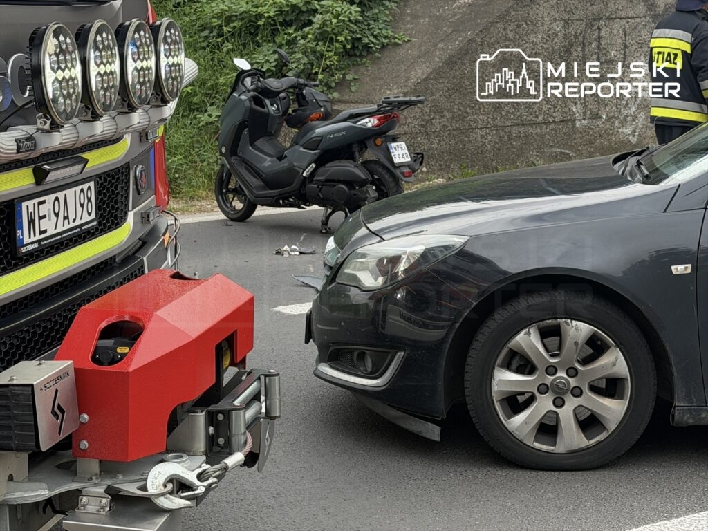 Czerwony pojazd straży pożarnej zatrzymany przy drodze, obok uszkodzony motocykl i czarny samochód osobowy.