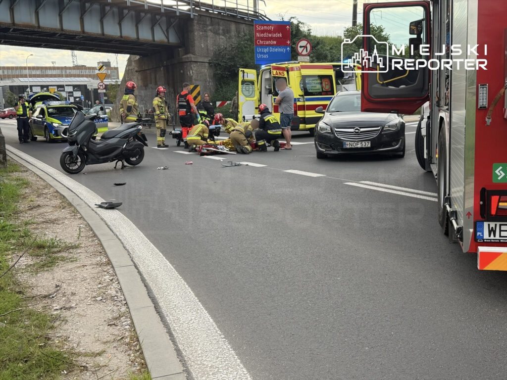 Grupa ratowników medycznych oraz strażaków udziela pomocy motocykliście na jezdni, obok uszkodzonego skutera.