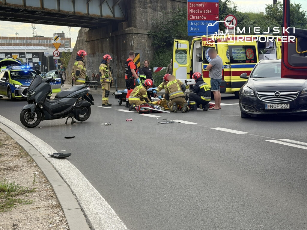 Motocyklista leży na jezdni, a ratownicy medyczni udzielają mu pomocy w pobliżu zderzenia z innym pojazdem.