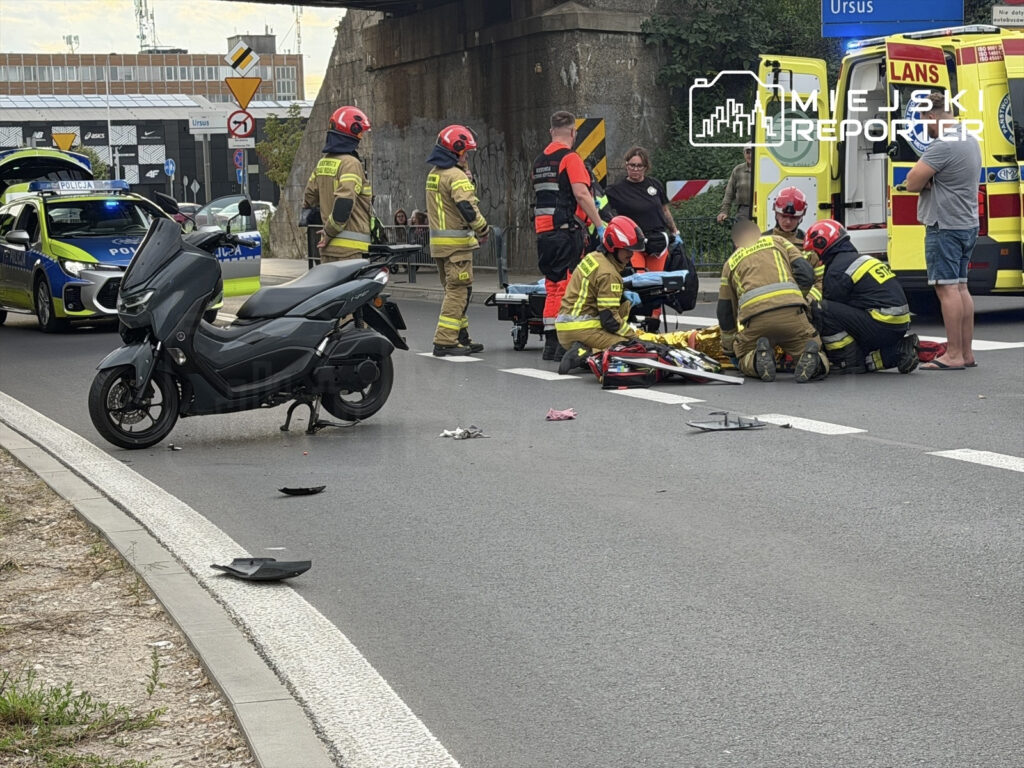 Motocyklista leży na jezdni, a zespół ratunkowy udziela mu pomocy, obok stoi uszkodzony skuter.