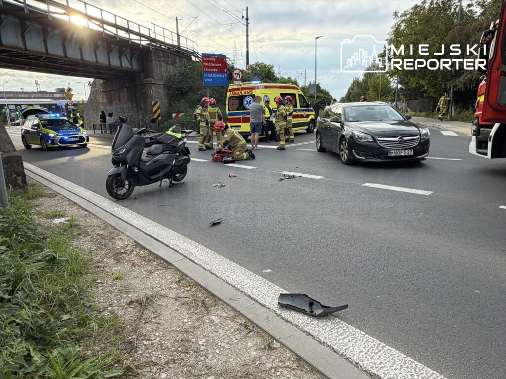 Motocyklista leży na jezdni obok uszkodzonego skutera, a w pobliżu pracują strażacy i policja.