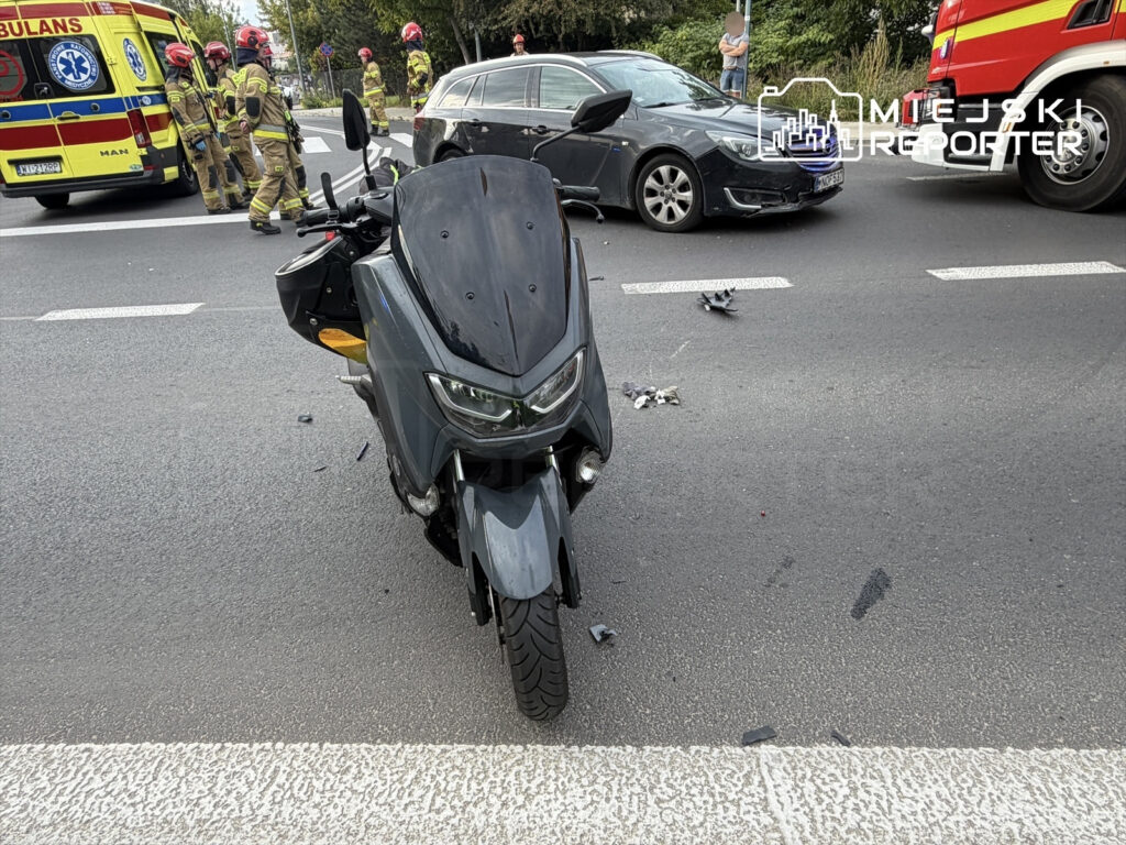Motocykl leżący na jezdni obok wypadku, w tle strażacy i karetka pogotowia. Wokół rozrzucone elementy pojazdu.