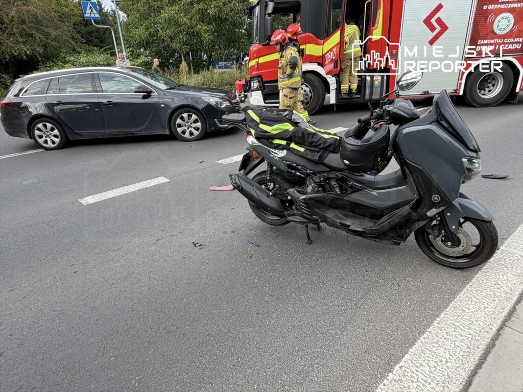 Motocyklista leżący obok uszkodzonego skutera na drodze, obok stoi czarny samochód osobowy oraz strażacy.