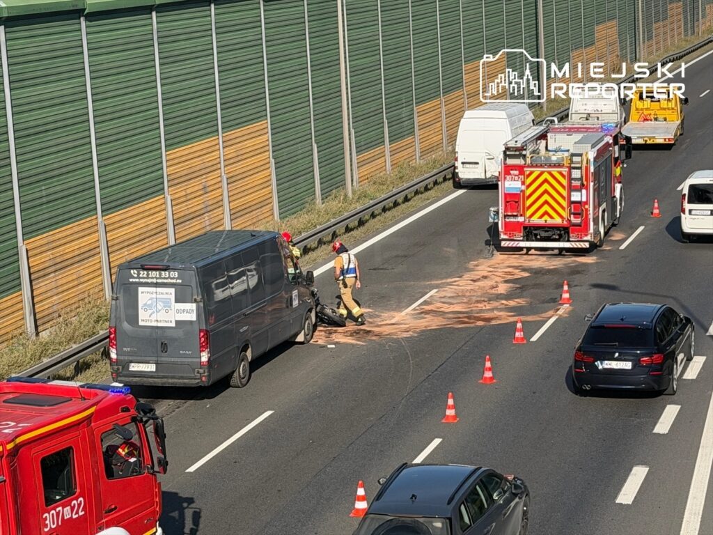 Motocyklista leży na jezdni obok busa, a strażacy oraz służby ratunkowe zabezpieczają miejsce wypadku.