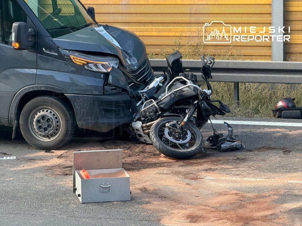Zniszczony motocykl leży na drodze obok uszkodzonego busa, a wokół rozrzucony jest piasek.