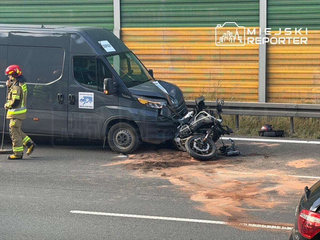 Motocyklista leży obok przewróconego motocykla na jezdni, obok stoi strażak w mundurze, a w tle widać szary bus.