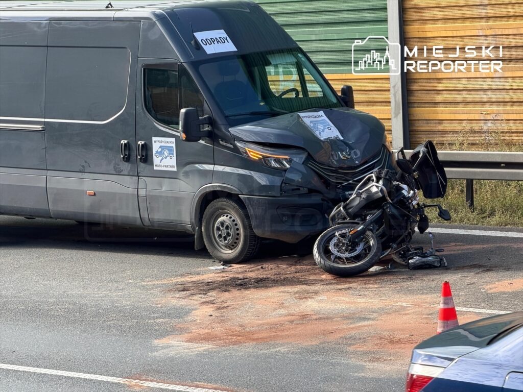 Motocykl leży na drodze obok szarego busa, na asfalcie widać plamy, a obok stoi czerwona pachołek.