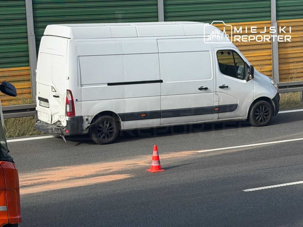 Biały bus z uszkodzeniami stoi na poboczu drogi, obok leży pomarańczowy proszek, a w tle widać zieloną barierkę.