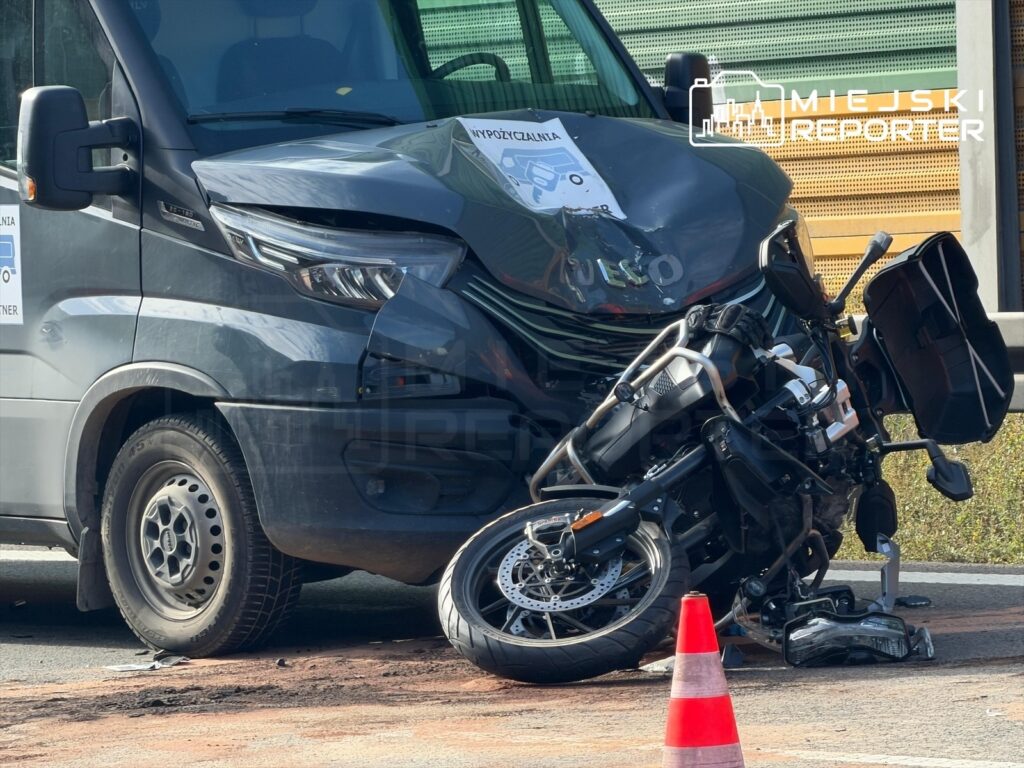 Zniszczony motocykl leży na jezdni obok zderzonego busa, z widocznymi uszkodzeniami nadwozia i leżącą obok czerwoną konusową zaporą.