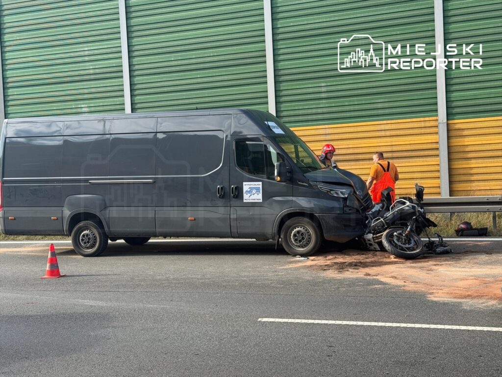 Motocyklista leży na drodze obok przewróconego motocykla, a obok niego stoi czarny bus z dwoma ratownikami.