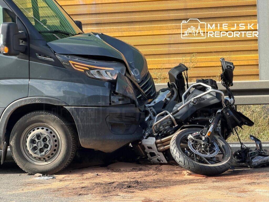 Zniszczony motocykl leży na drodze obok uszkodzonego busa, na asfalcie widoczne plamy oleju.