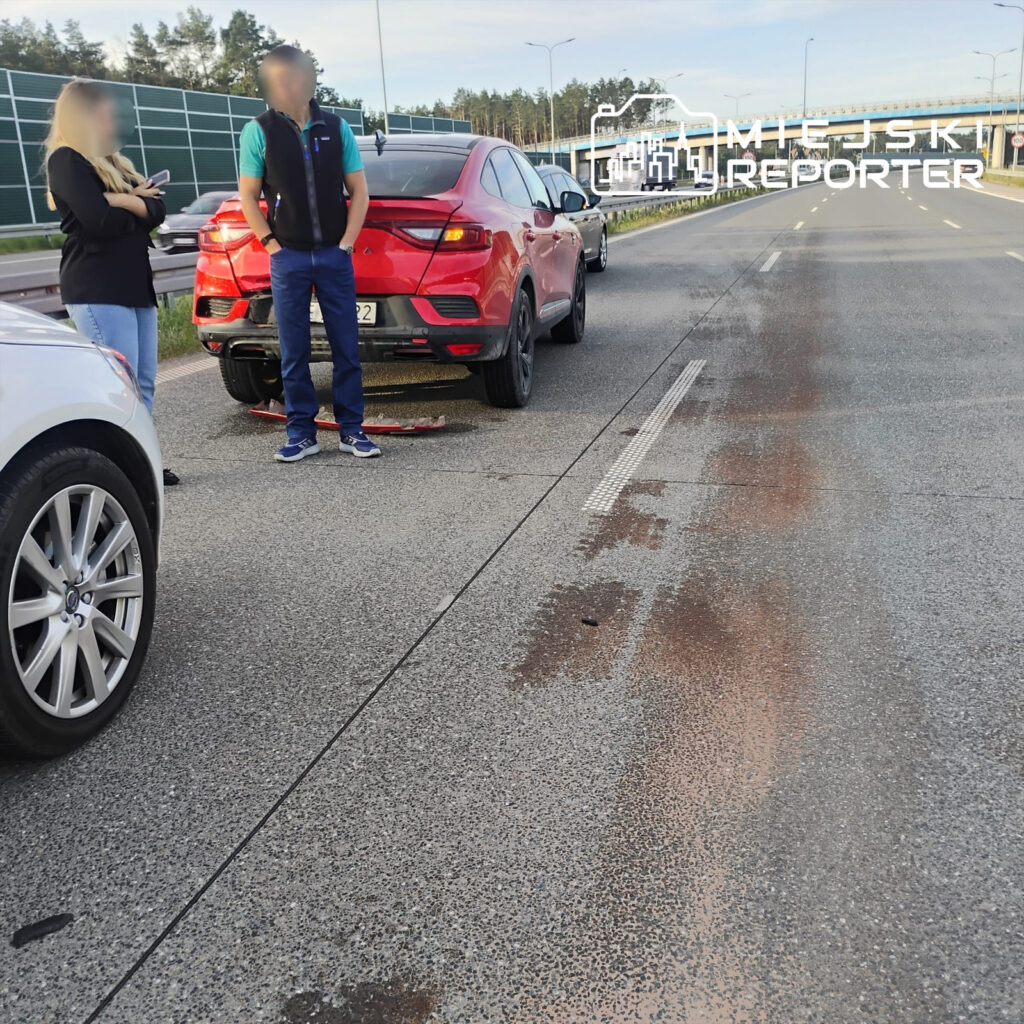 Kobieta w czarnej bluzce i mężczyzna w niebieskiej koszulce stoją obok uszkodzonego czerwonego SUV-a na drodze.