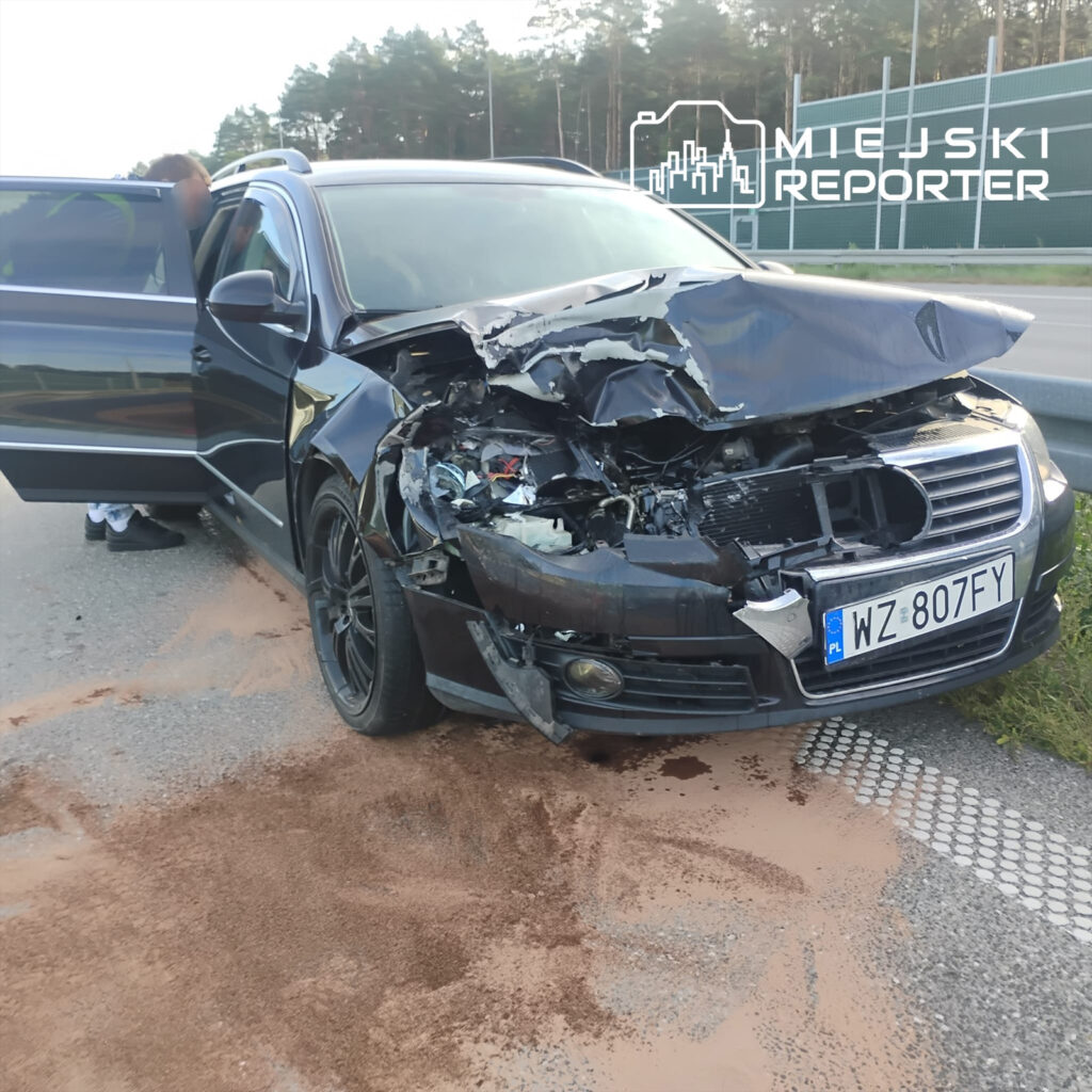 Czarny samochód osobowy z uszkodzonym przodem stoi na drodze, obok niego mężczyzna rozmawia z kierowcą.