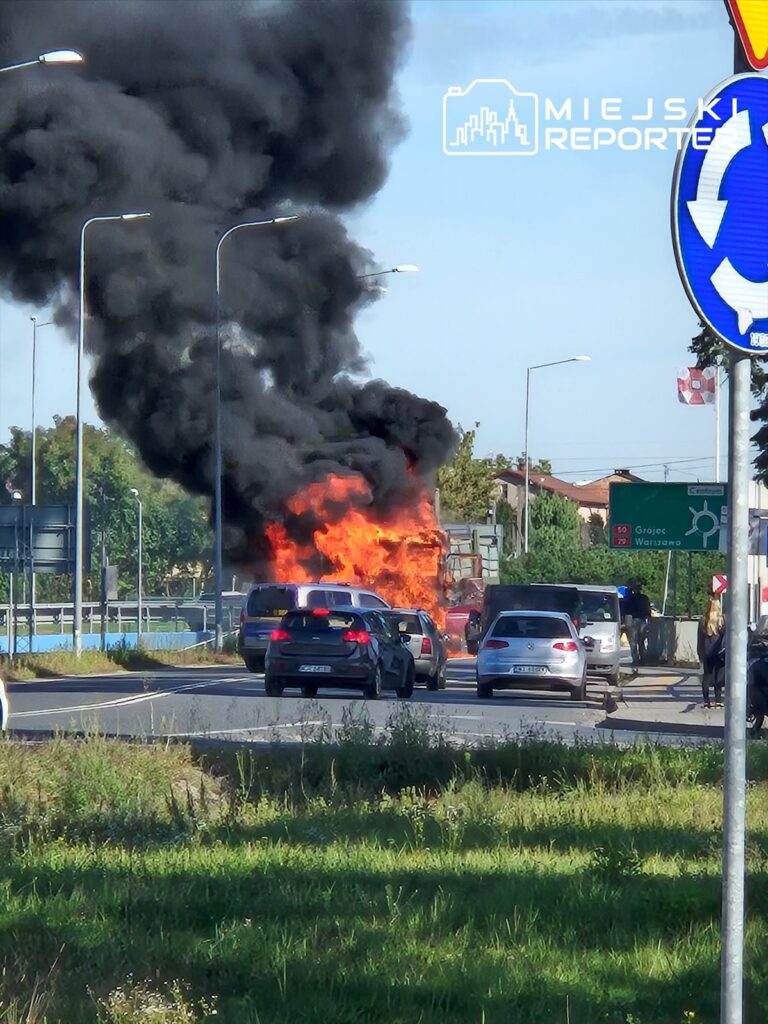 Ciężarówka w płomieniach wydobywa czarny dym na ruchliwej drodze, obok stoją zatrzymane samochody.