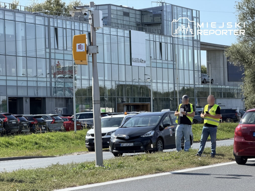 Dwaj mężczyźni w odblaskowych kamizelkach sprawdzają fotoradar obok nowoczesnego biurowca z przeszkloną fasadą.
