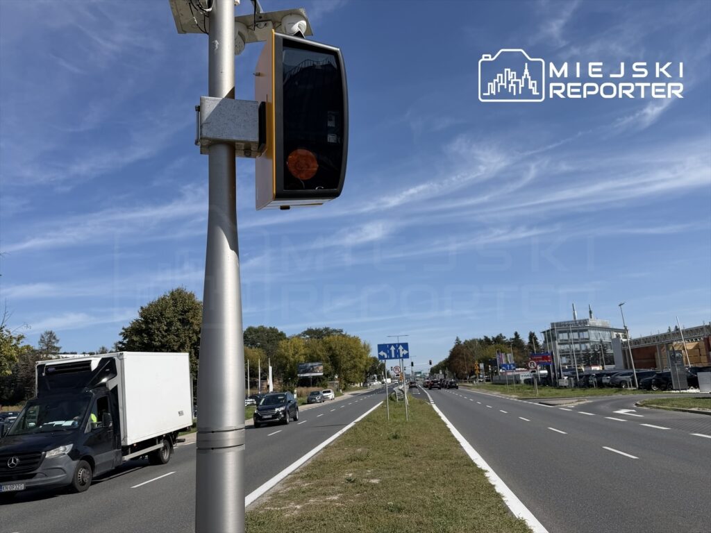 Fotoradar zamontowany na słupie, monitorujący ruch na dwupasmowej drodze w słoneczny dzień.