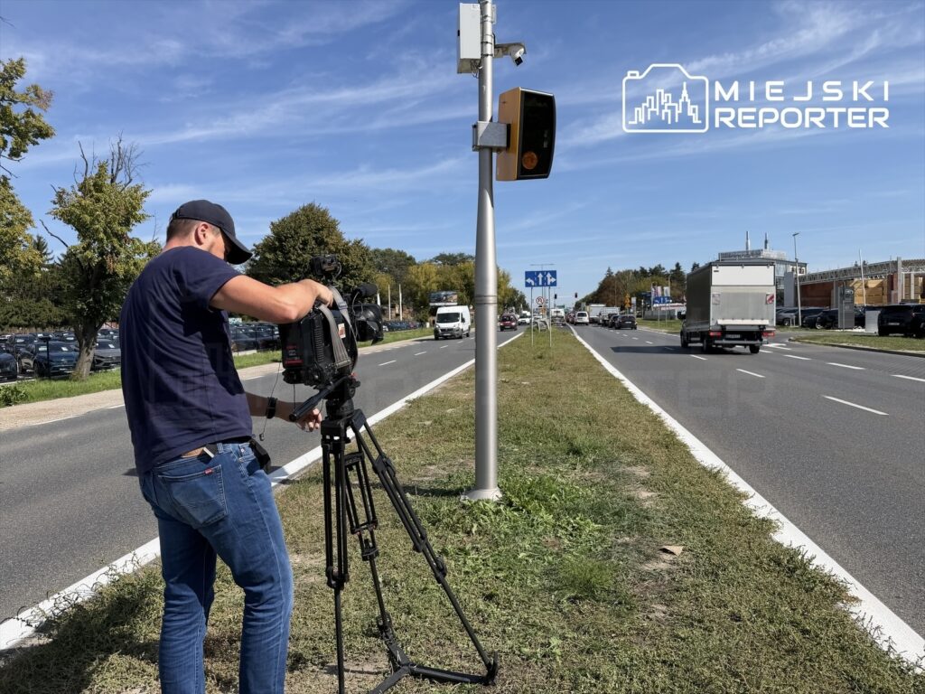 Mężczyzna ustawia kamerę na statywie obok ruchliwej drogi, w tle widać fotoradar oraz przejeżdżające pojazdy.