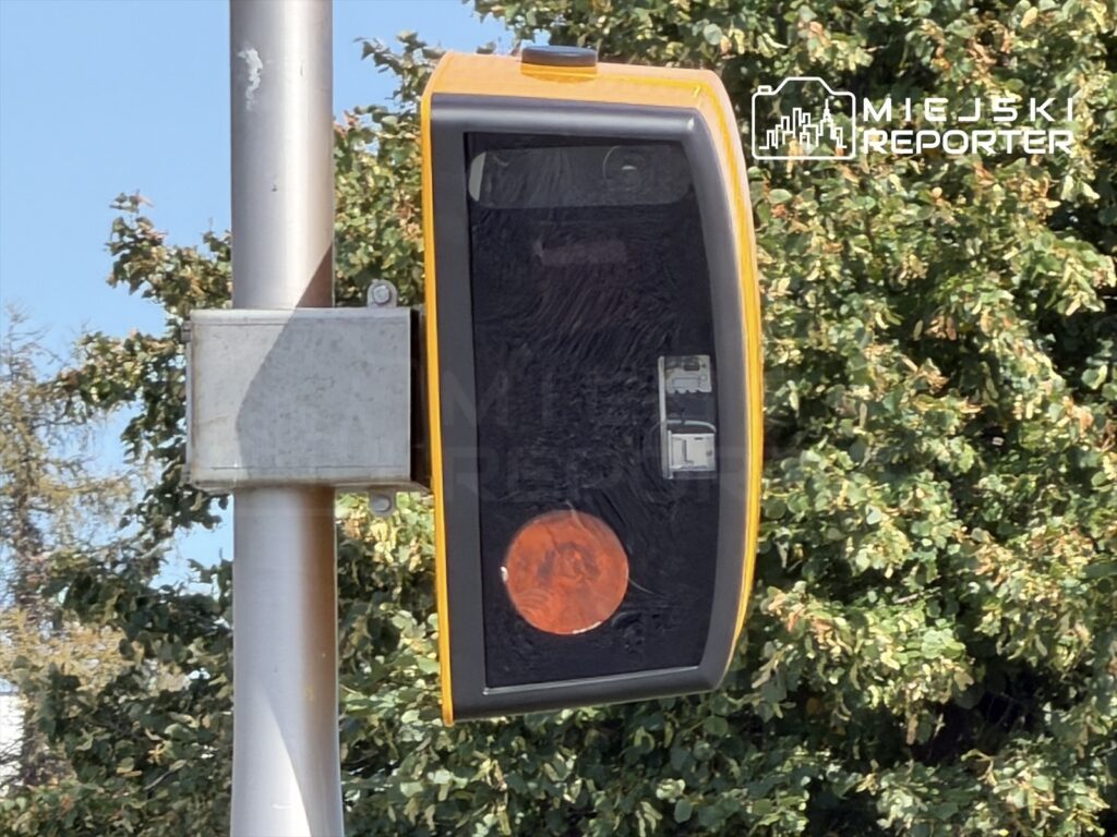 Fotoradar zamontowany na słupie, z widocznym czerwonym sygnalizatorem, w tle zieleń drzew.