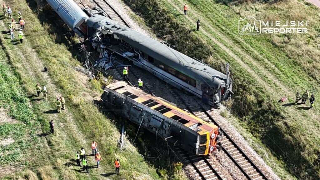 Wielu ratowników pracuje przy przewróconym pociągu na torach, obok uszkodzonego wagonu i zniszczeń.