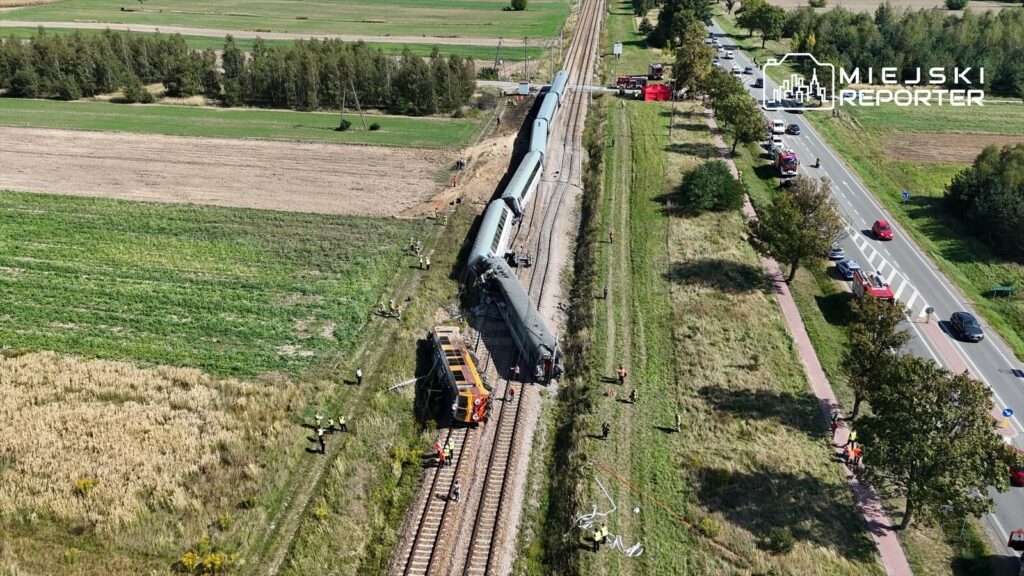Wieloczłonowy pociąg wykolejony obok torów, w tle widać zespół ratunkowy oraz samochody na drodze.